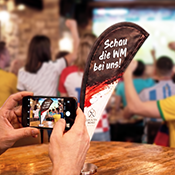 Hände einer Person, die mit ihrem Smartphone eine Tischfahne im WM-Design fotografiert, im Hintergrund sind jubelnde Menschen beim Public Viewing mit Deutschland-Fanartikeln zu sehen Hände einer Person, die mit ihrem Smartphone eine Tischfahne im WM-Design fotografiert, im Hintergrund sind jubelnde Menschen beim Public Viewing mit Deutschland-Fanartikeln zu sehen