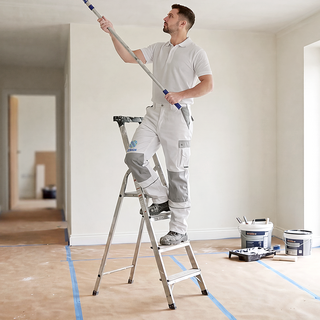 Ein Mann in bestickter Basic-Arbeitshose steht auf einer Leiter und streicht eine Zimmerdecke
